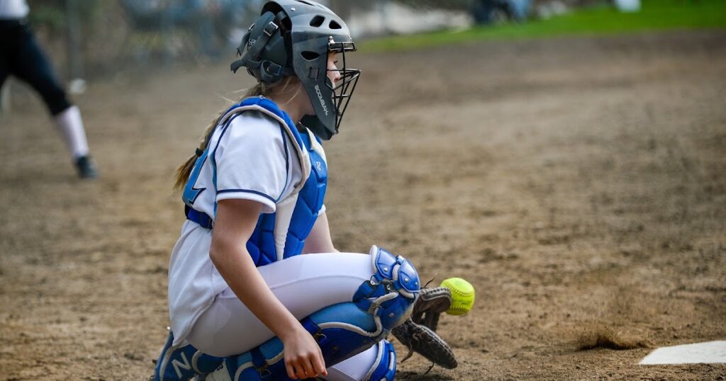 A girl in catcher's gear kneels to catch a ball, embodying the spirit of the Best Softball Team Names in action.